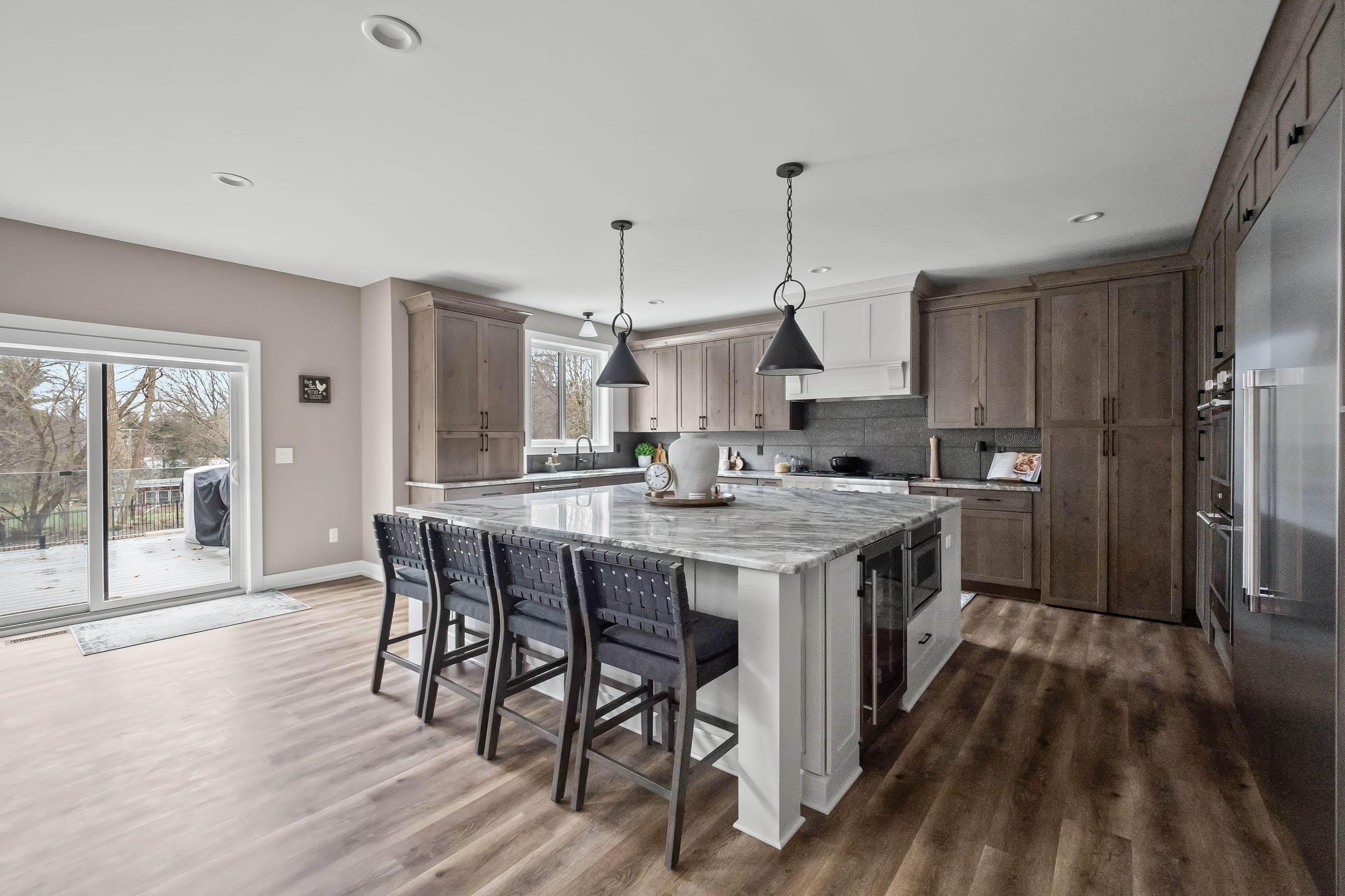 Open-concept view of the kitchen showcasing the large island with seating, Rustic Alder cabinetry, white hood, and sliding doors to the outdoor deck.