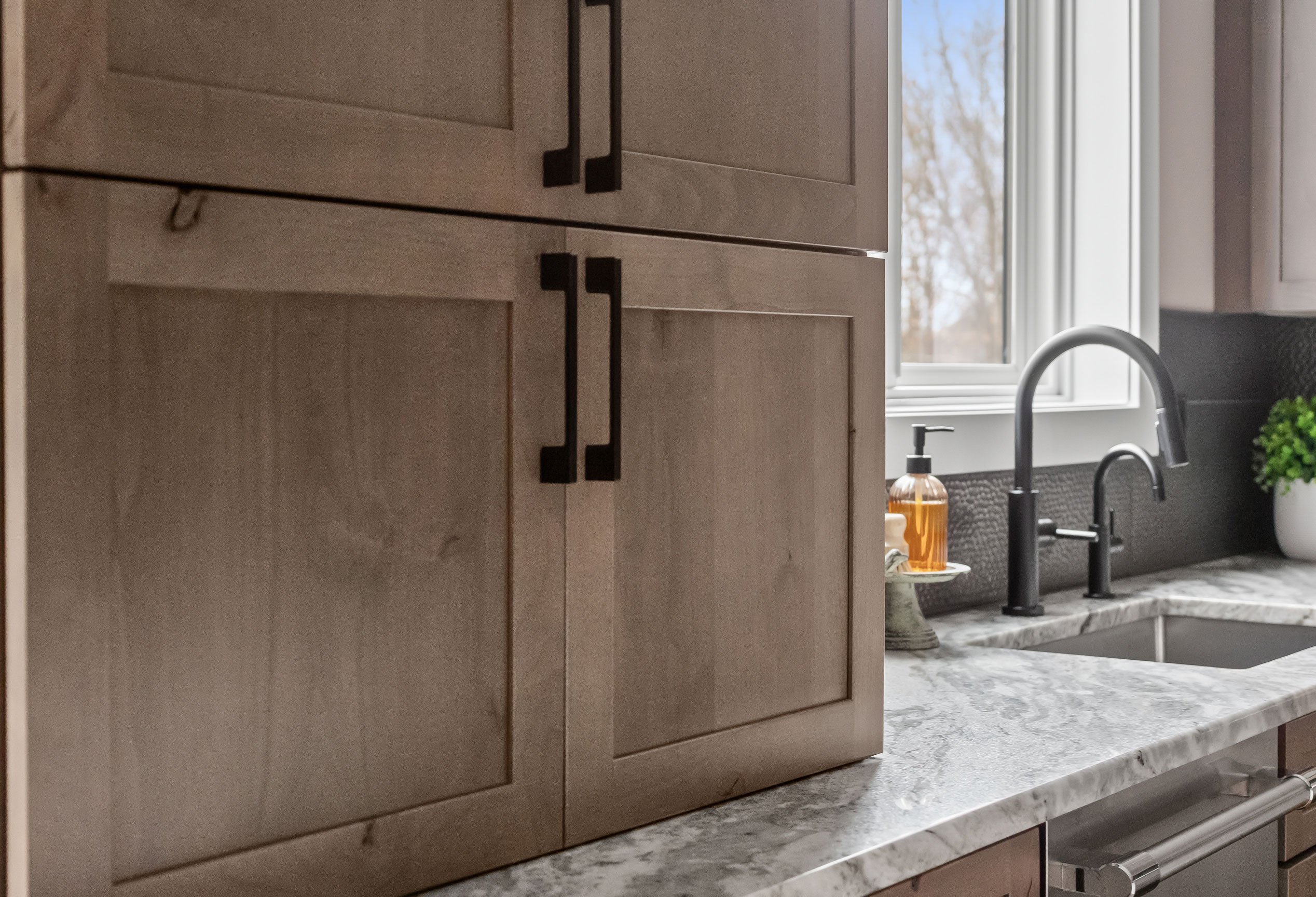 Kitchen sink area featuring Rustic Alder cabinetry appliance garage, matte black faucet, and Fantasy Brown granite countertops under a large window.