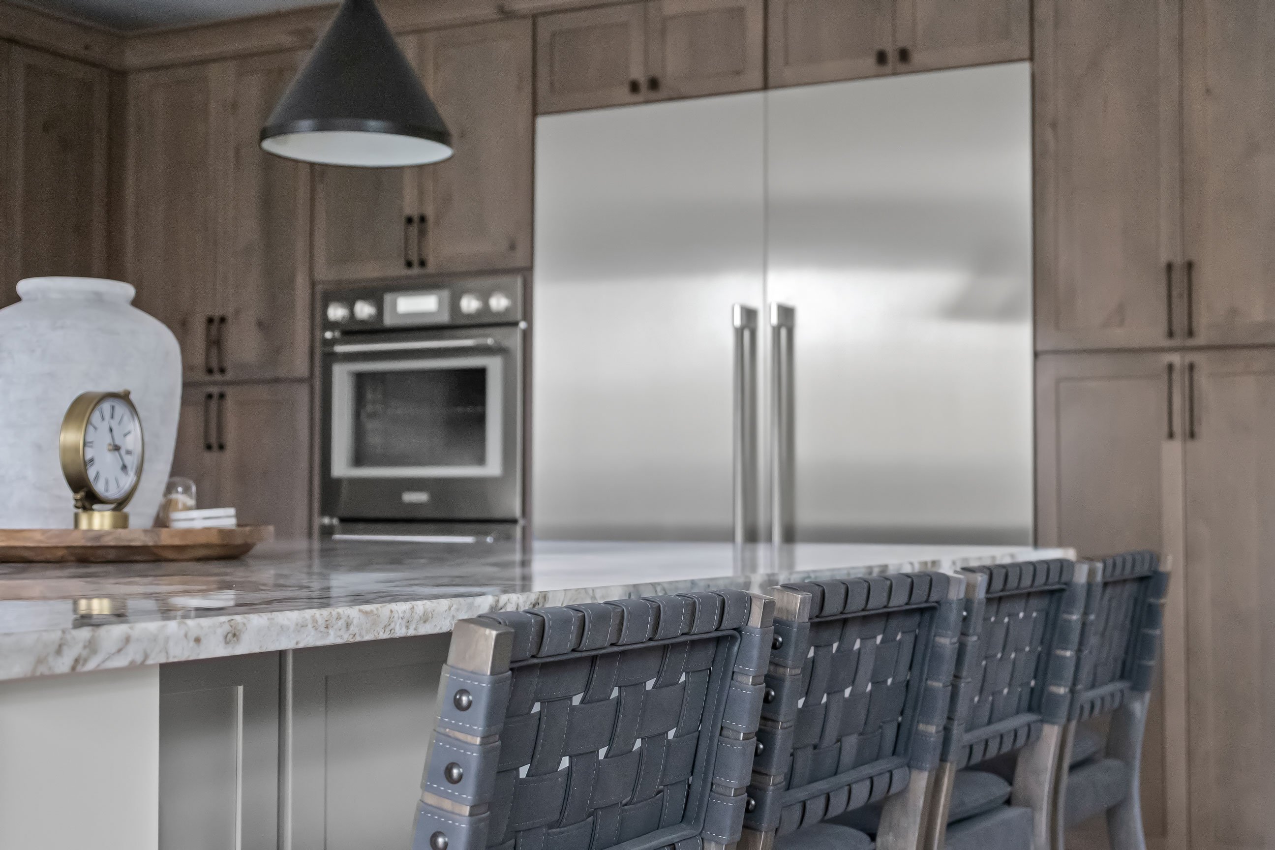 Large built-in refrigerator and double wall ovens integrated into Rustic Alder cabinetry, viewed from behind the island’s woven-back bar stools.
