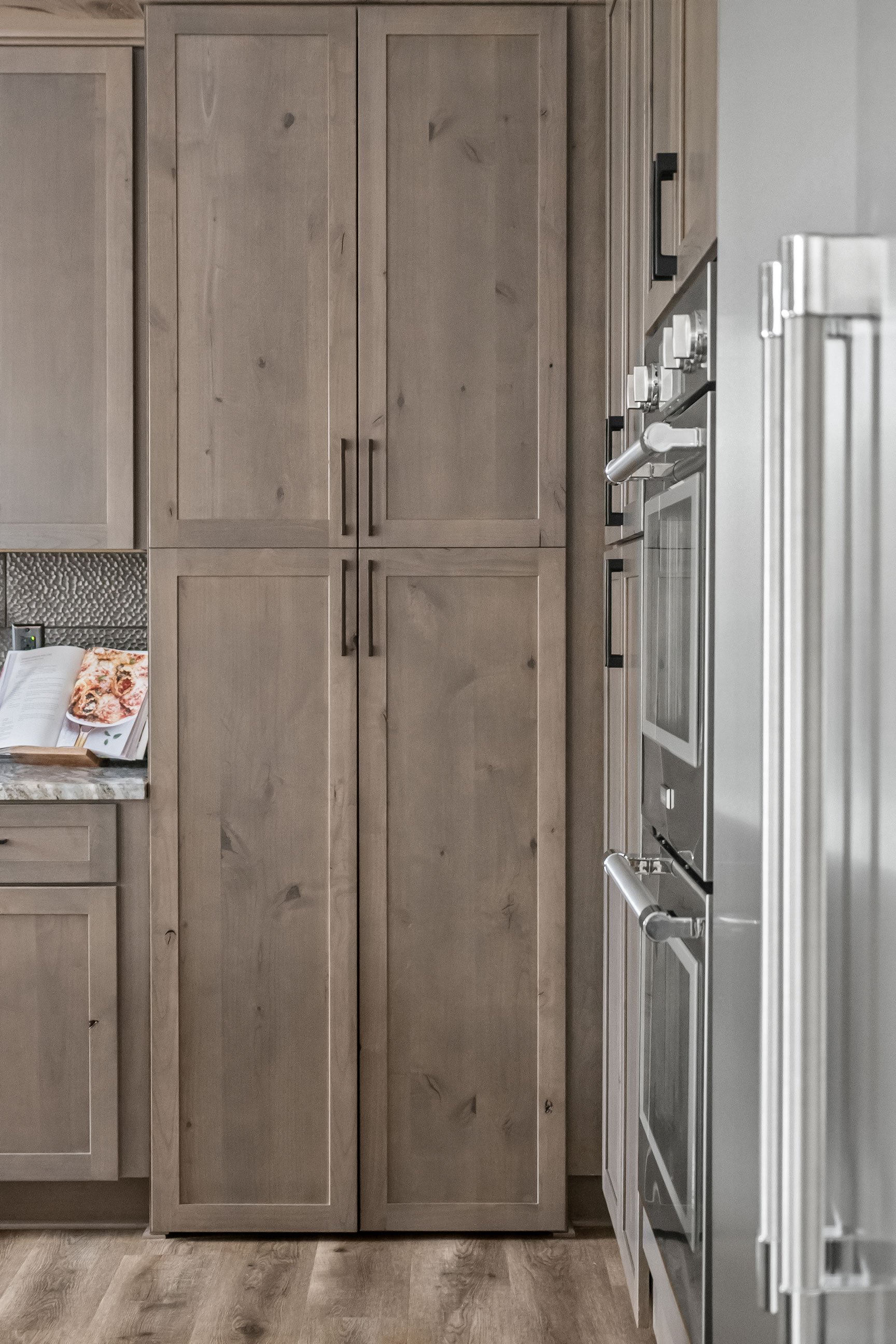 Rustic Alder tall cabinetry concealing walk-through pantry, adjacent to stainless steel double ovens and a large built-in refrigerator, creating a seamless wall of storage.