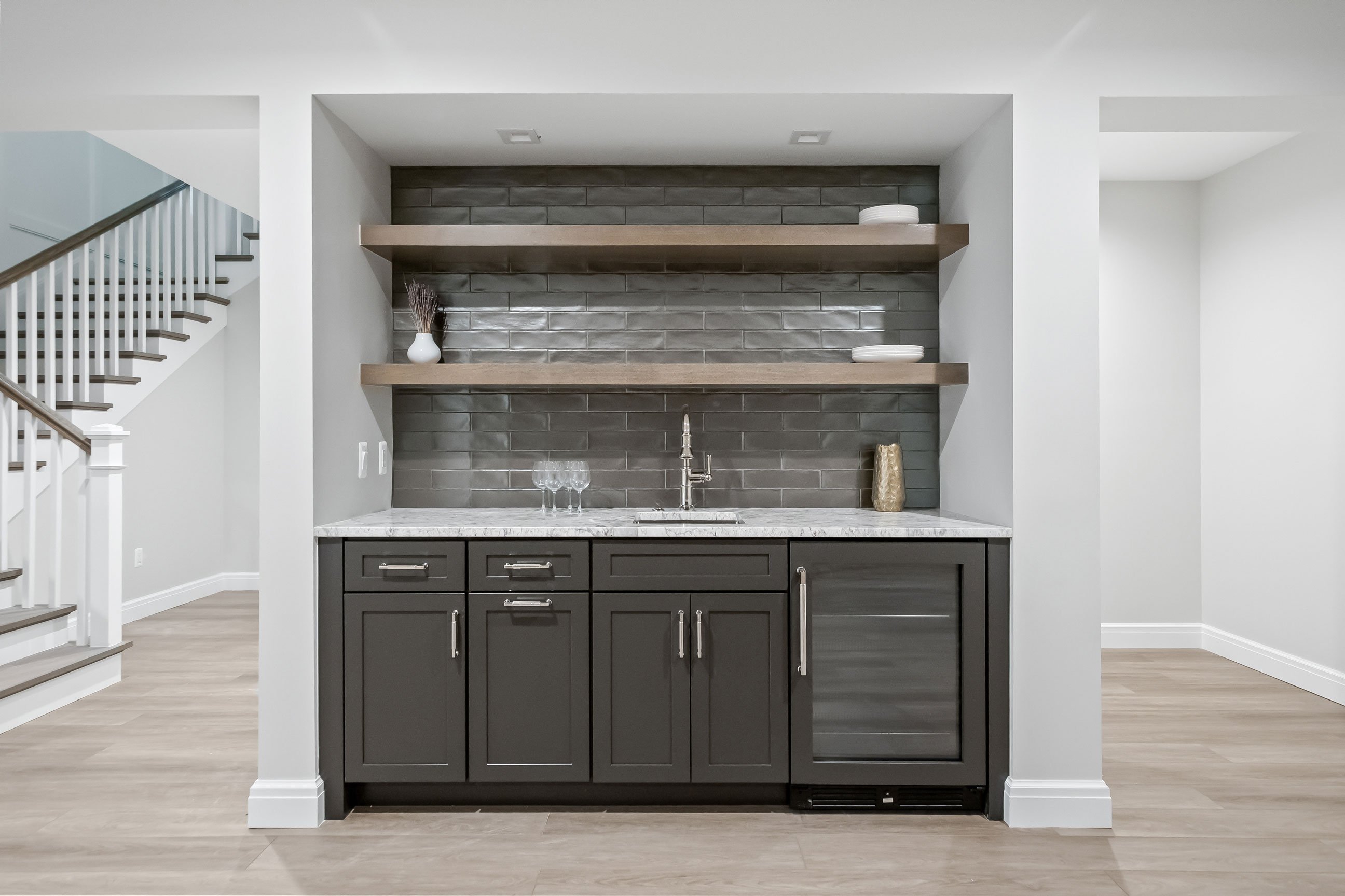 Lower-level bar area with moody dark cabinetry, gray tile backsplash, and floating shelves designed by KSI for entertaining in style.