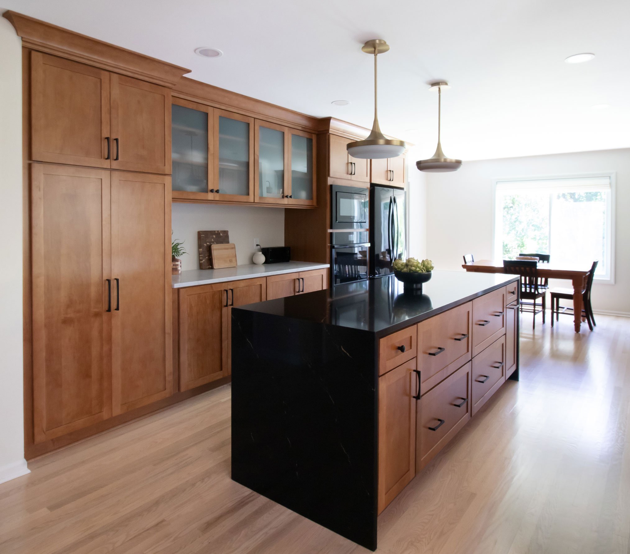 Warm kitchen remodel featuring shaker cabinetry with warm wood stain and black accents, with black quartz waterfall countertop on island - view showing island, pantry, and food prep area