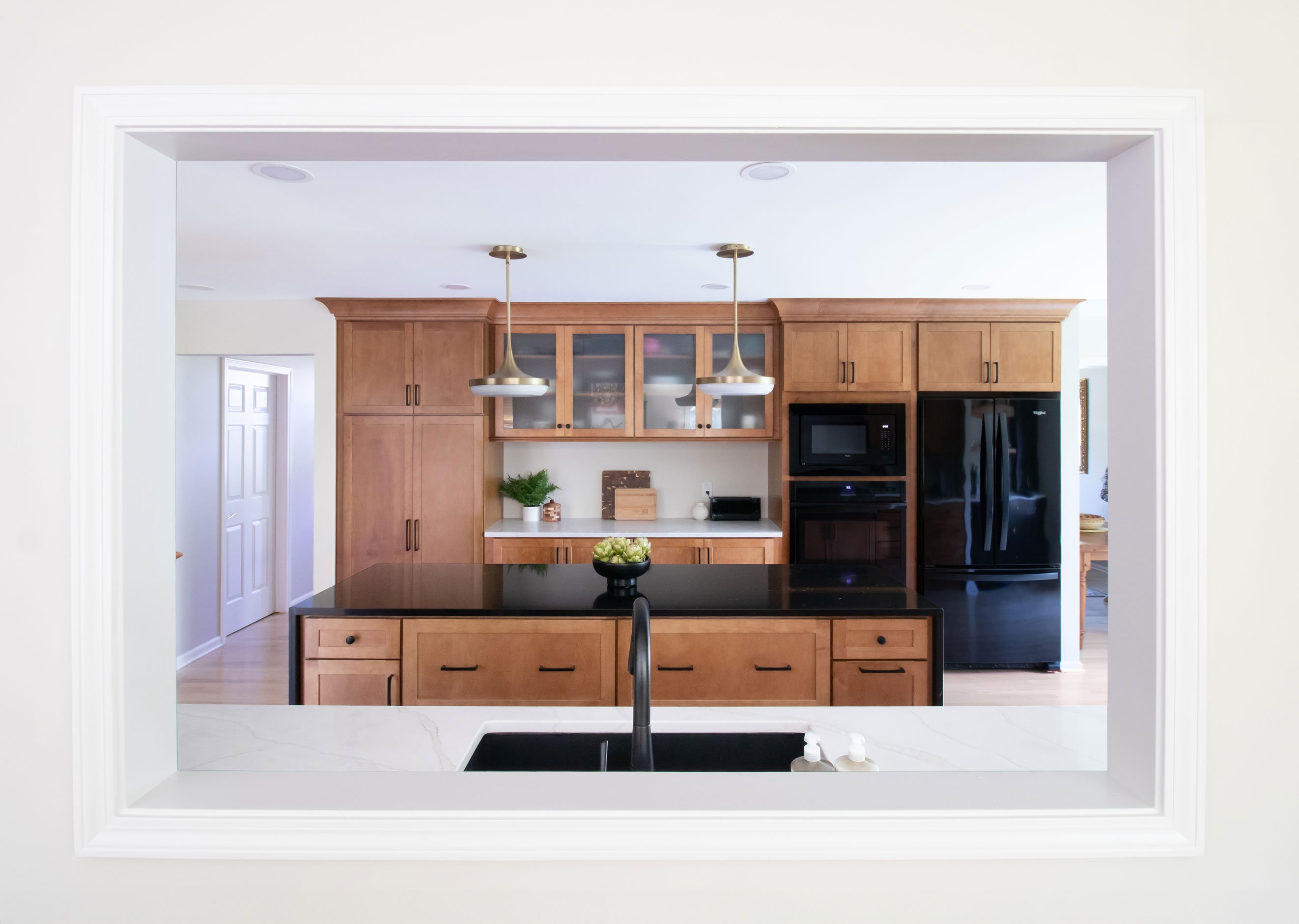 Warm kitchen remodel featuring shaker cabinetry with warm wood stain and black accents, with black quartz waterfall countertop on island - view through living room pass-through showing island, pantry, and food prep area