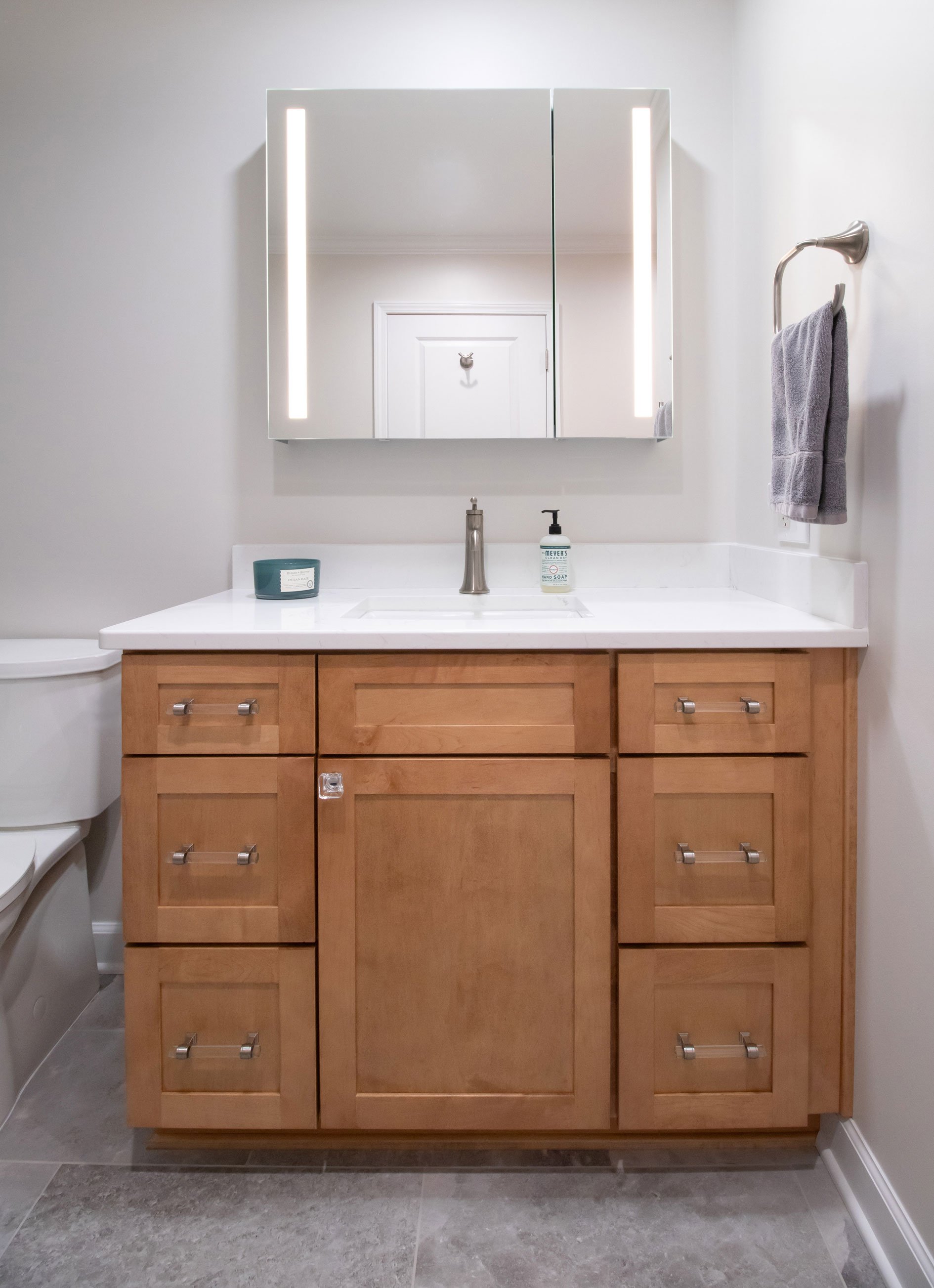Bath vanity with warm wood shaker cabinetry, Amerock designer hardware, quartz top, and lit mirror
