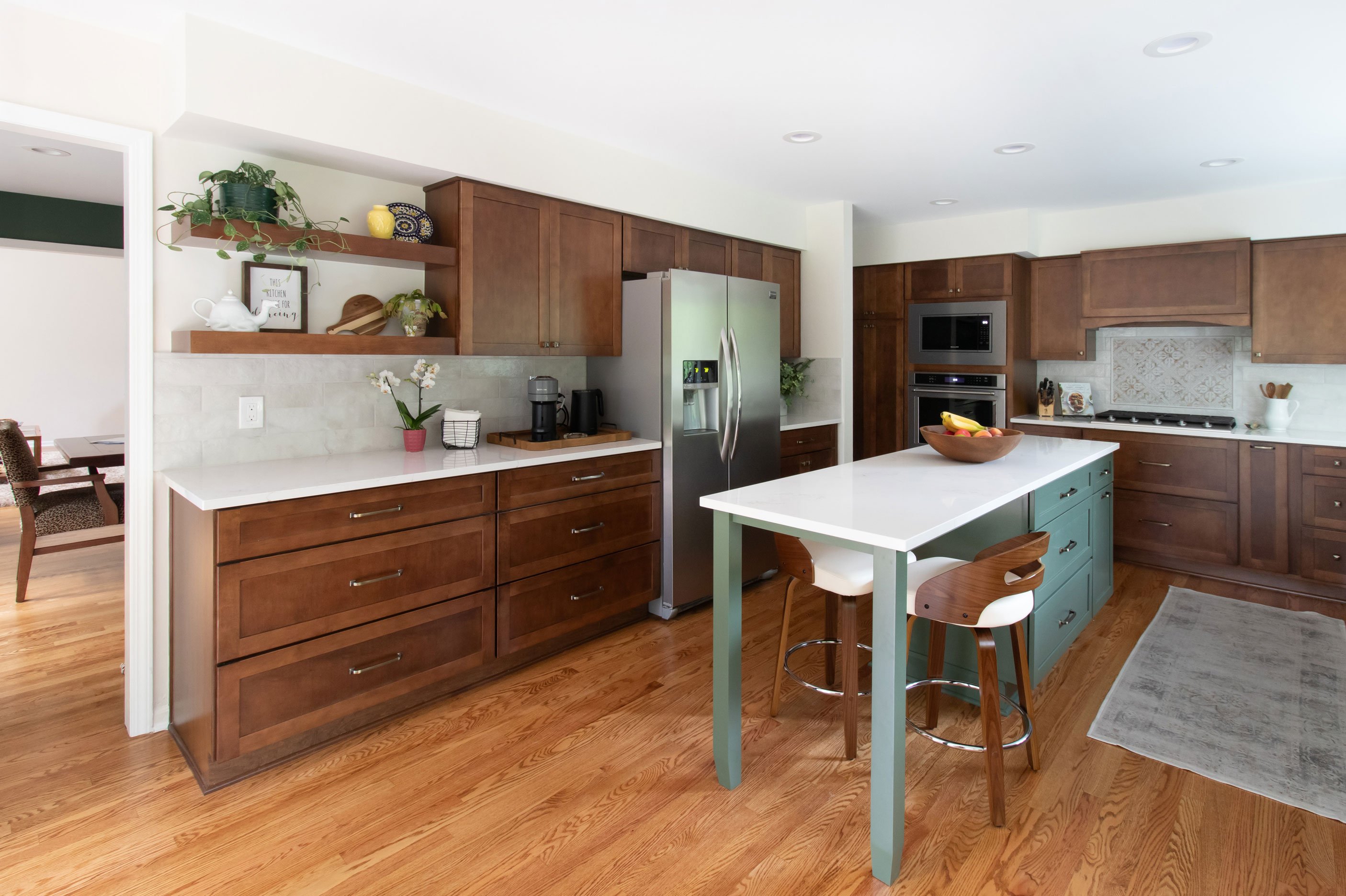 Wide view of remodeled kitchen with Merillat Masterpiece cabinetry in the Martel door style, featuring maple wood in Cognac finish on the perimeter and Evergreen paint on the island, and white quartz countertops.