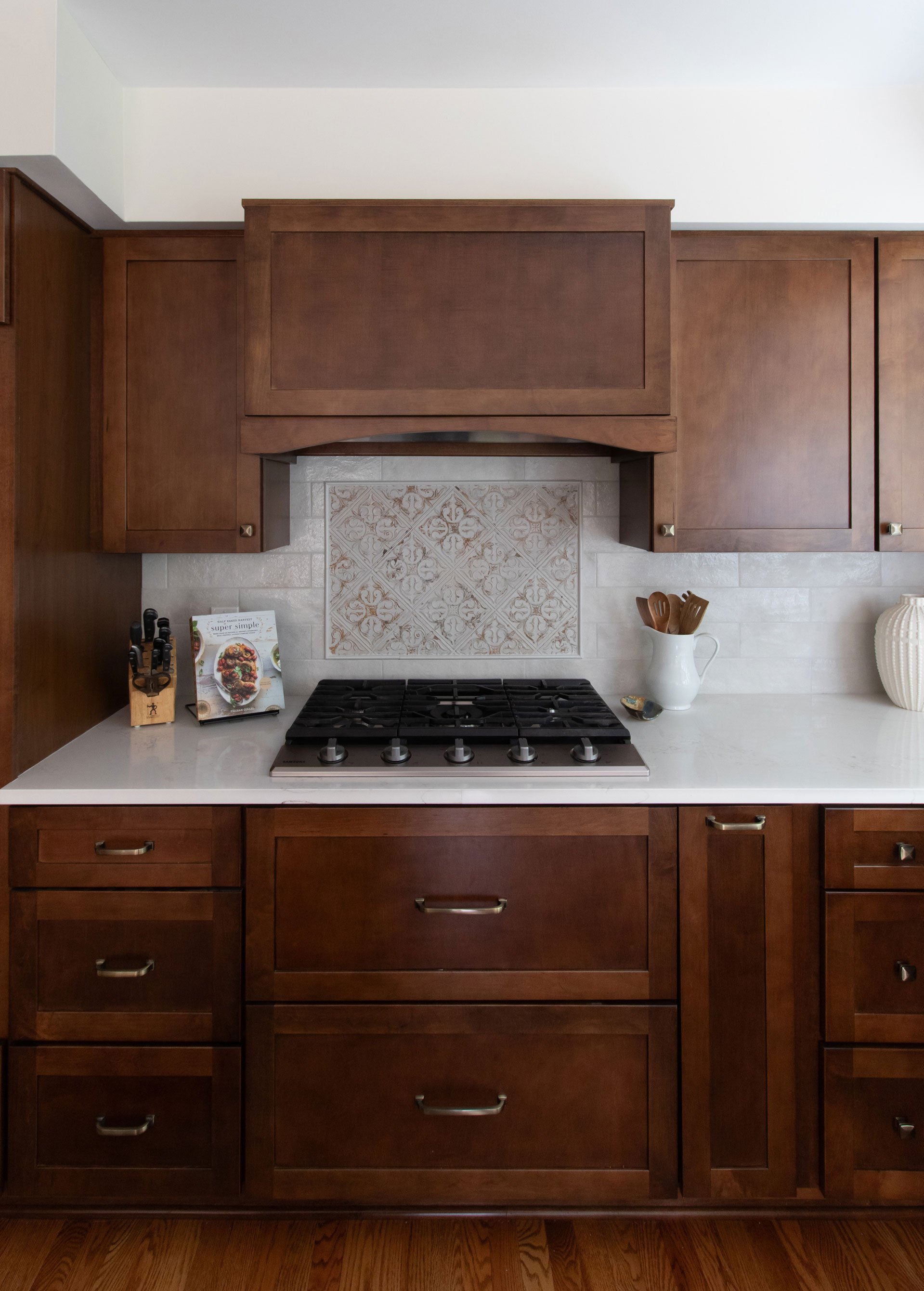 Cooktop wall with Merillat Masterpiece cabinetry in the Martel door style, featuring maple wood in Cognac finish, quartz countertop, and patterned backsplash accent.