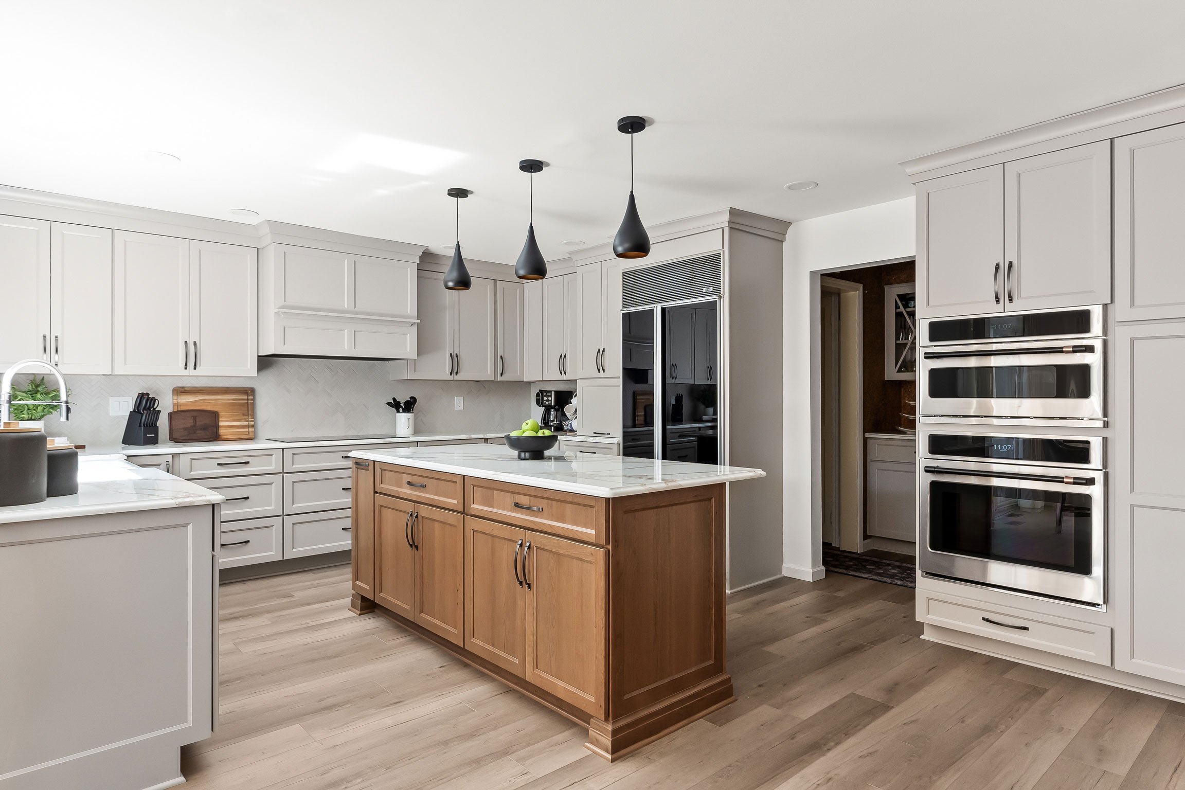 A dramatic kitchen remodel featuring warm greige perimeter cabinetry, a stained cherry island, black teardrop pendant lights, and a stainless steel Café wall oven tower.
