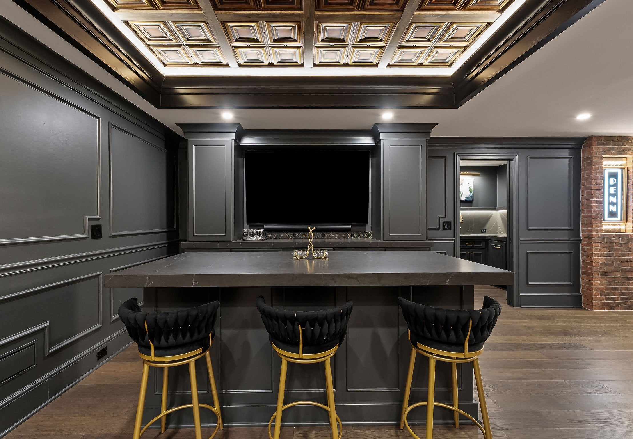 Speakeasy-inspired basement bar with charcoal cabinetry, large entertaining island, coffered illuminated ceiling, and built-in media wall.