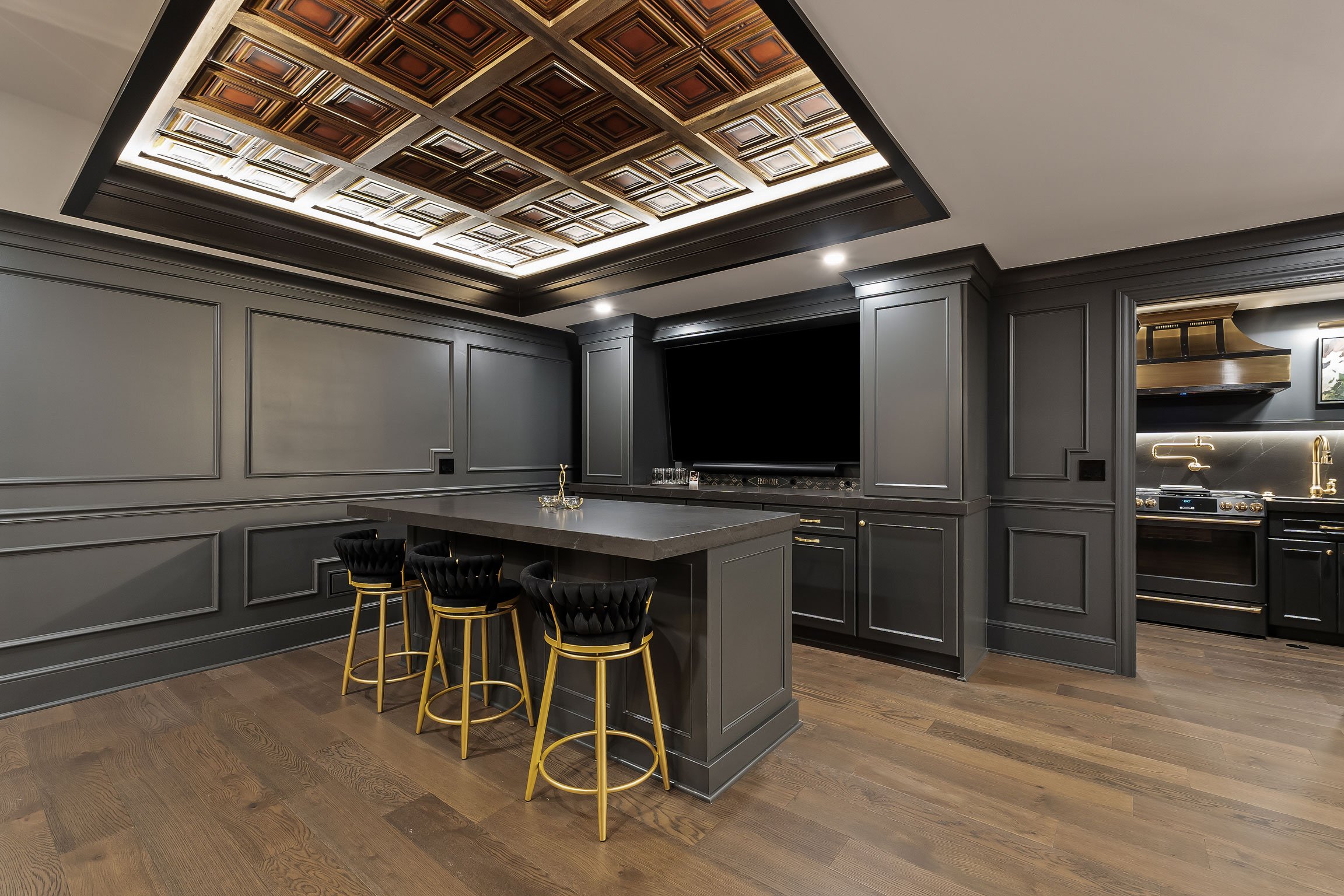 Luxury basement renovation showcasing illuminated coffered ceiling, custom bar island, secondary kitchen, and dramatic dark cabinetry.
