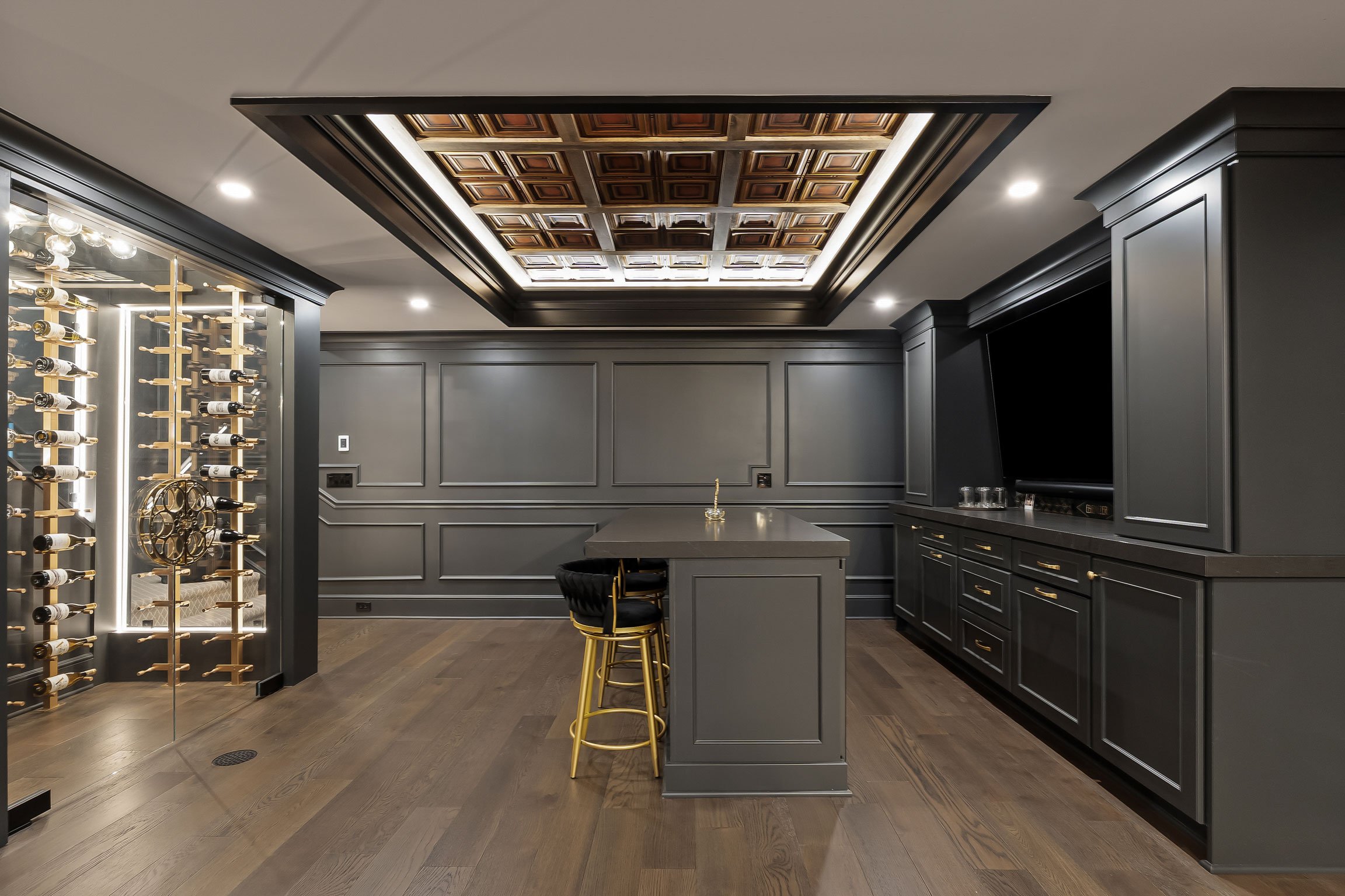 Custom basement entertaining space featuring a dark moody bar, glass-enclosed wine room, large island, and detailed wall molding.
