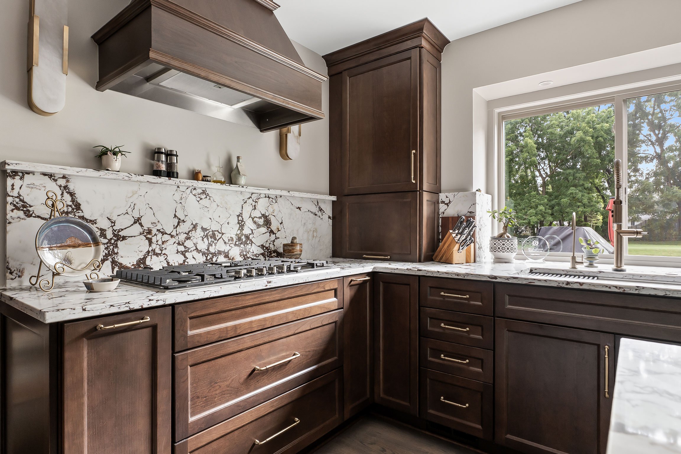 Close view of the wood range hood and stone backsplash above the cooktop, with a tall cherry cabinet and window to the side.