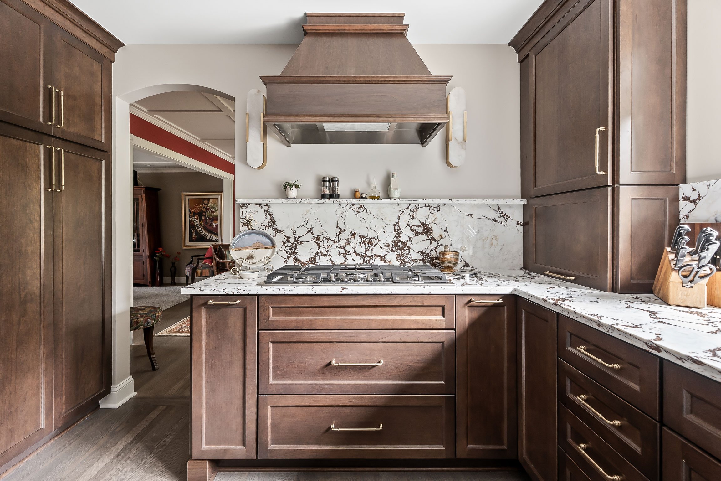 The kitchen's range wall viewed from the island, featuring a custom wood hood, veined stone backsplash, and cherry cabinetry with a glimpse into the adjoining living room.