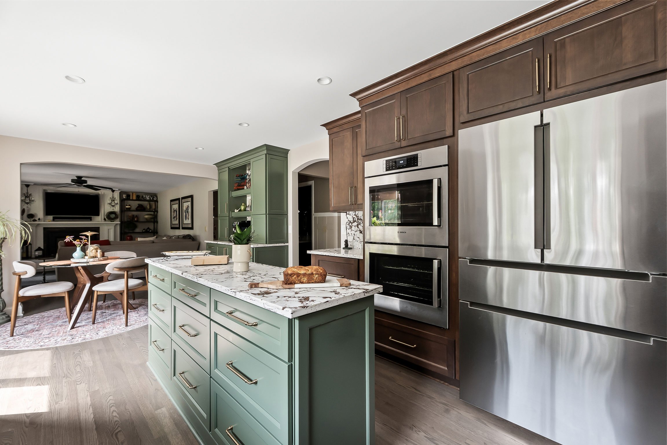 The refrigerator and double wall oven wall in Brown Sugar cherry cabinetry, with the green island and breakfast nook visible in the foreground.
