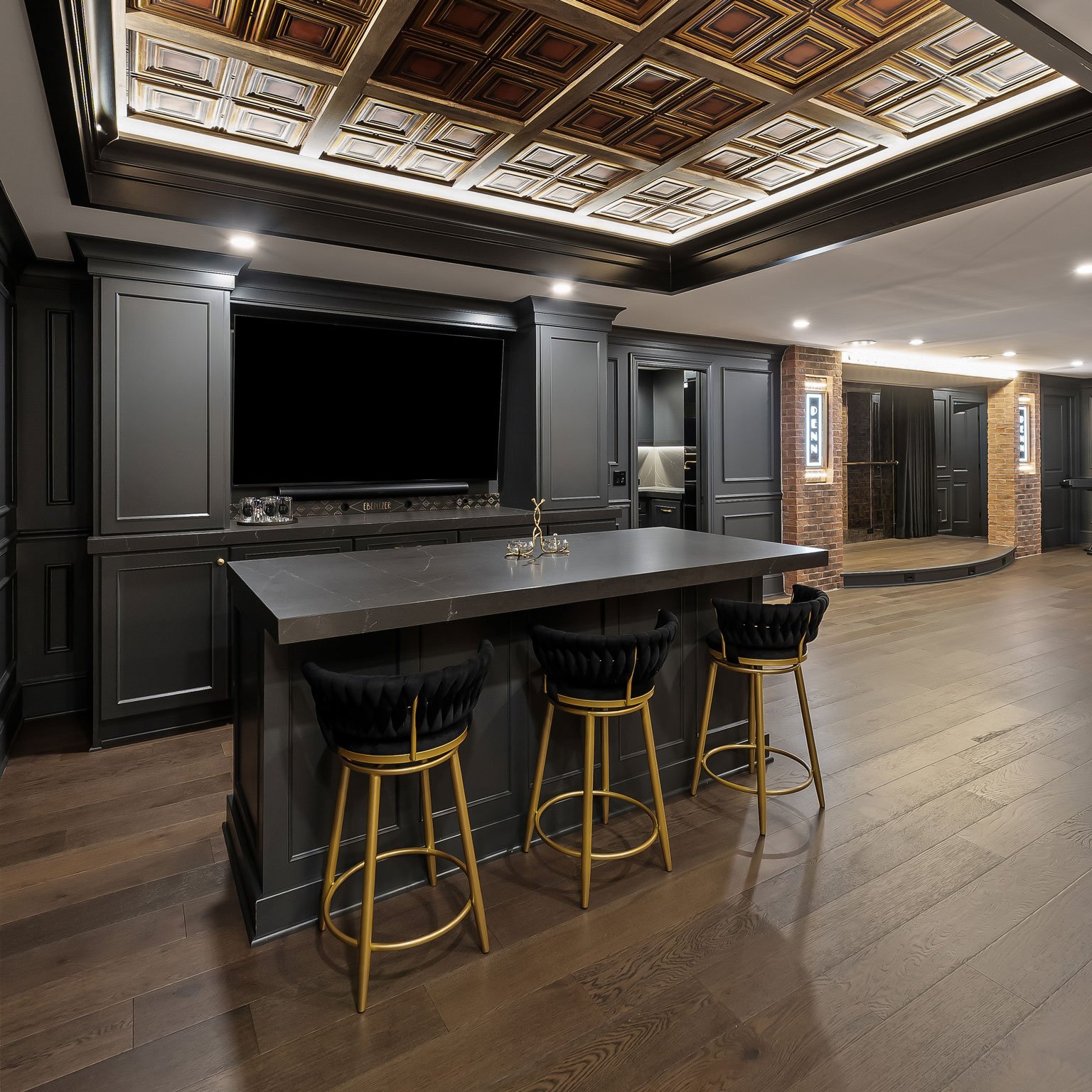 Luxury basement renovation showcasing illuminated coffered ceiling, custom bar island, theater stage, and dramatic dark gray cabinetry.