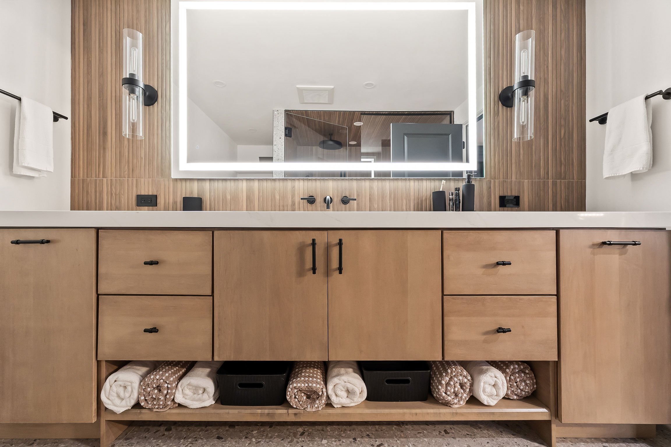 Contemporary basement bathroom featuring a warm wood vanity, illuminated mirror, and modern black fixtures.