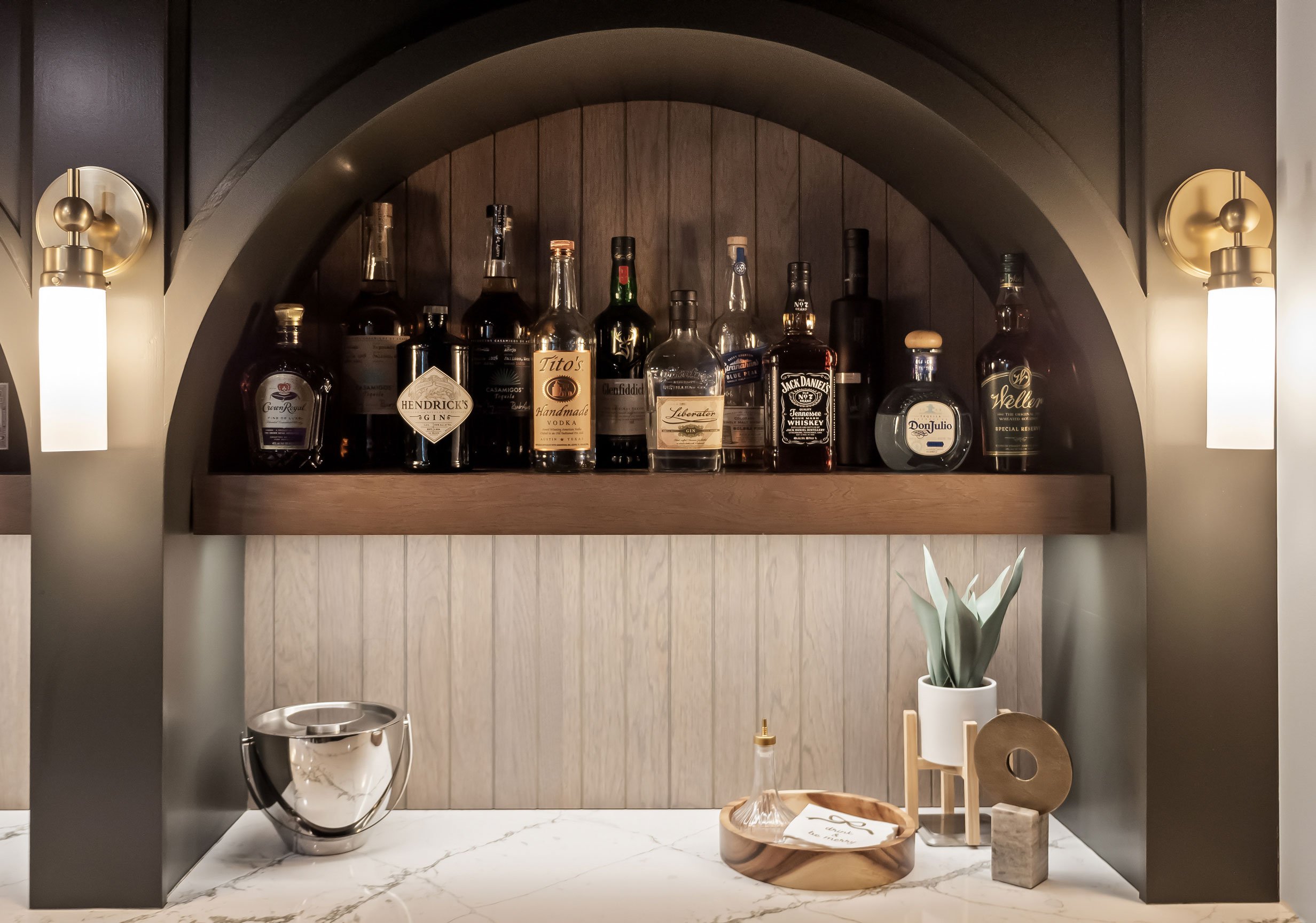 Speakeasy-style basement bar detail with arched detailing, floating wood shelves, dark cabinetry, quartz countertops, and warm accent lighting.