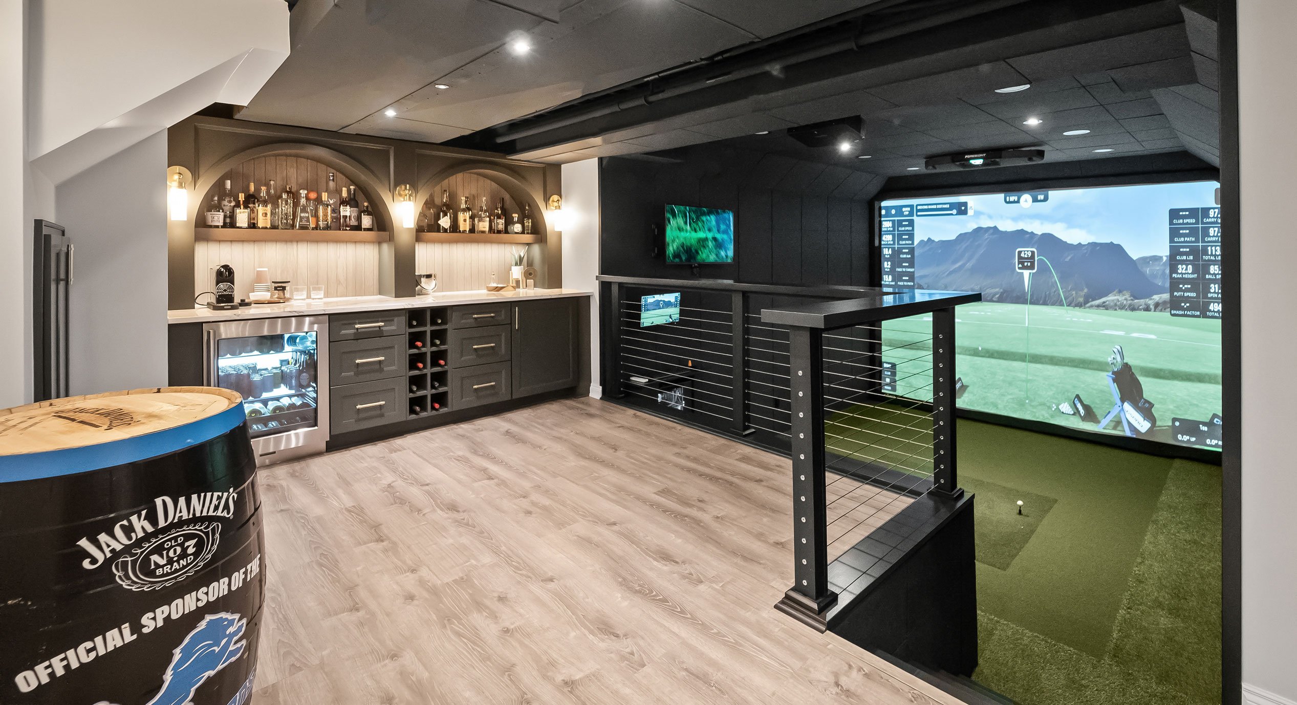 Golf simulator in a finished basement with open railing overlook connected to a kitchen and bar entertainment space