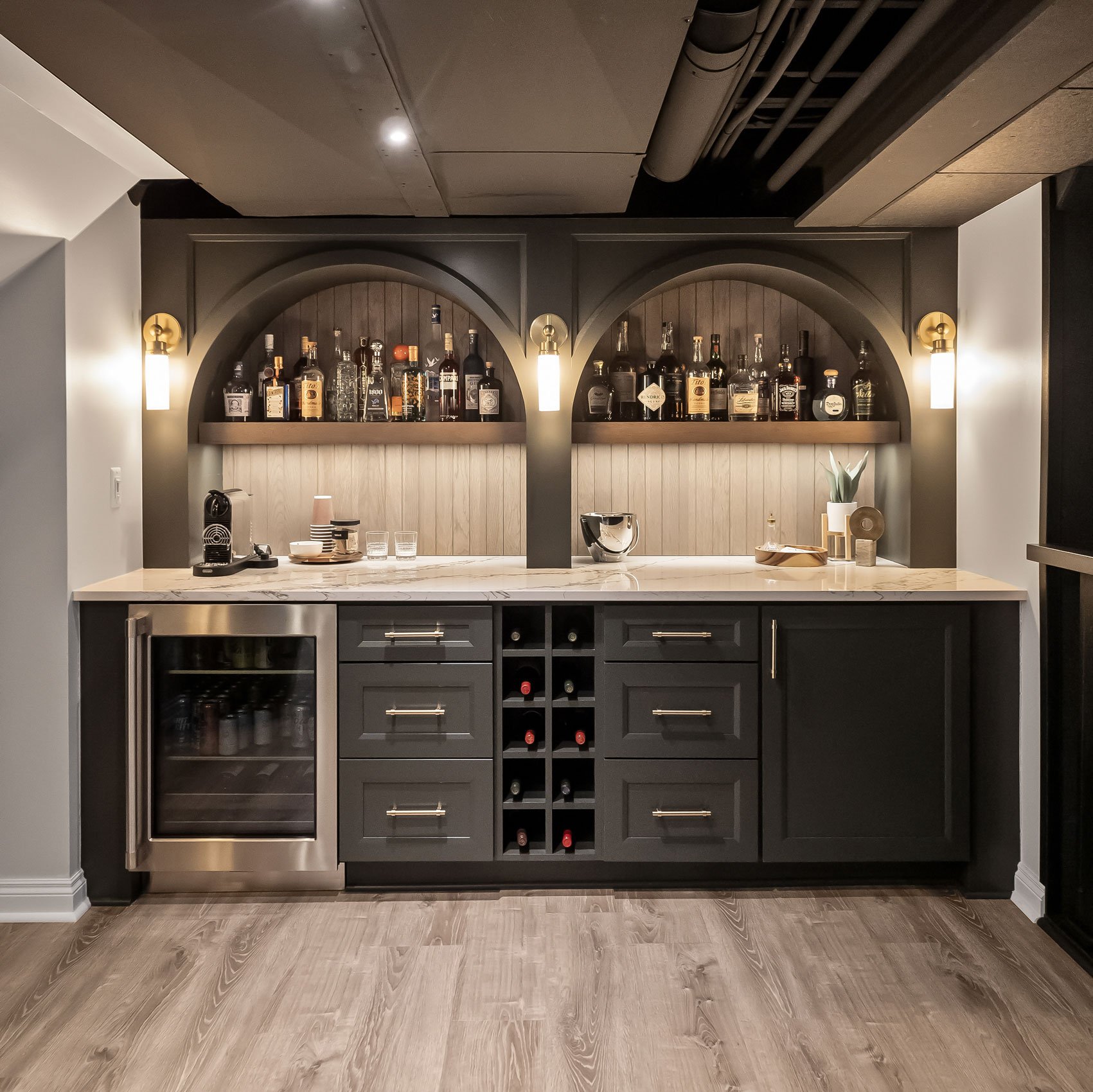 Speakeasy-style basement bar with arched detailing, floating wood shelves, dark cabinetry, quartz countertops, and warm accent lighting.