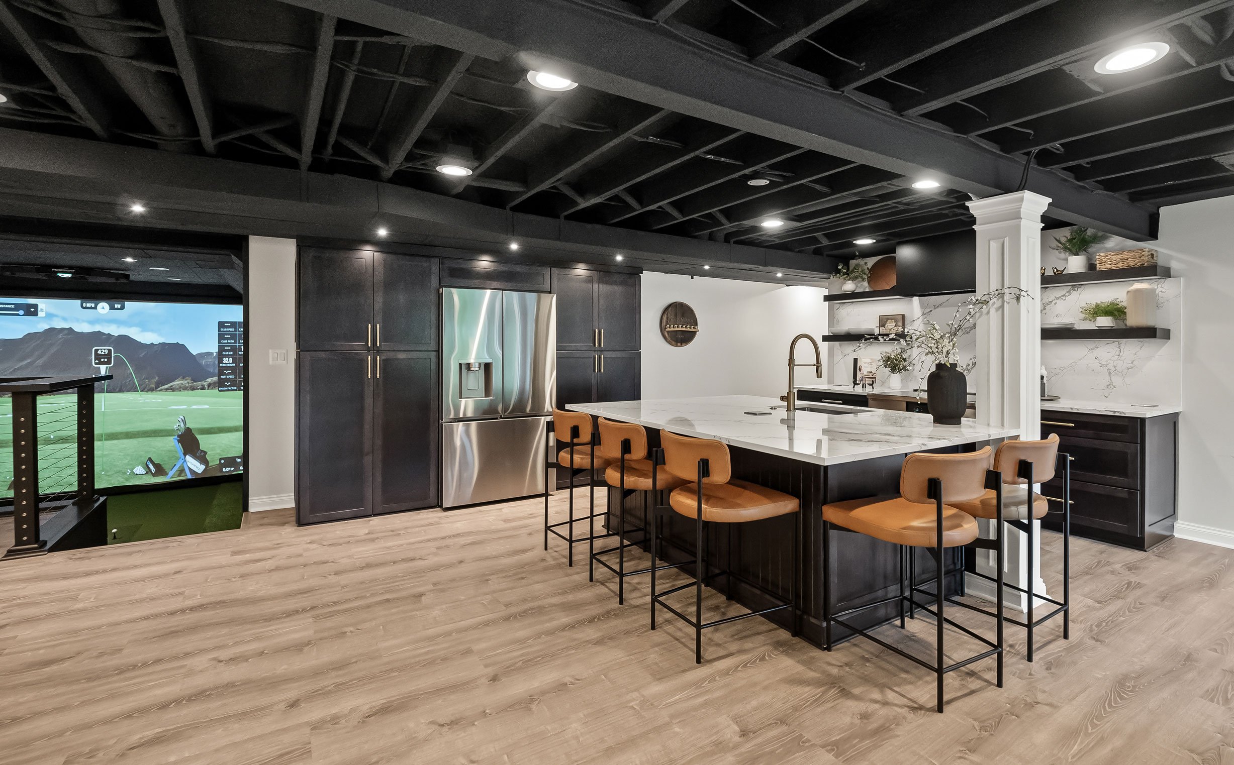 Basement entertainment kitchen with dark cabinetry, large island seating, quartz countertops, and open layout overlooking a golf simulator.
