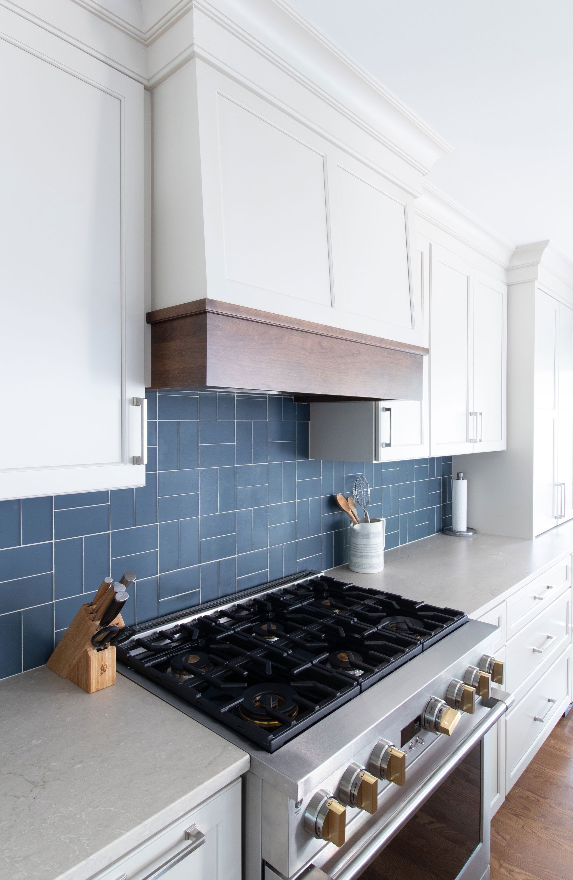 Professional-style gas range beneath a custom wood hood with blue tile backsplash and white shaker cabinetry.