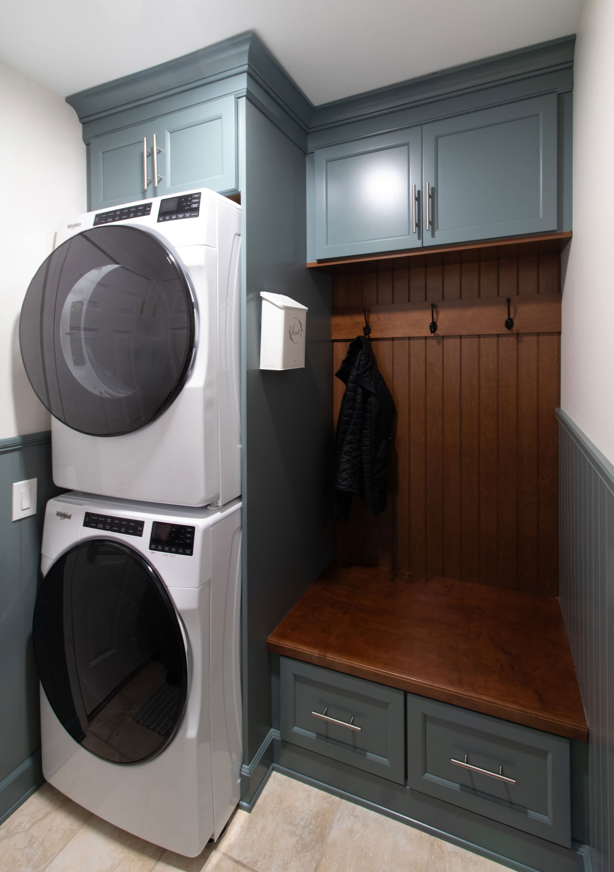 Custom laundry room with stacked washer and dryer, built-in bench, storage drawers, coat hooks, and blue cabinetry.