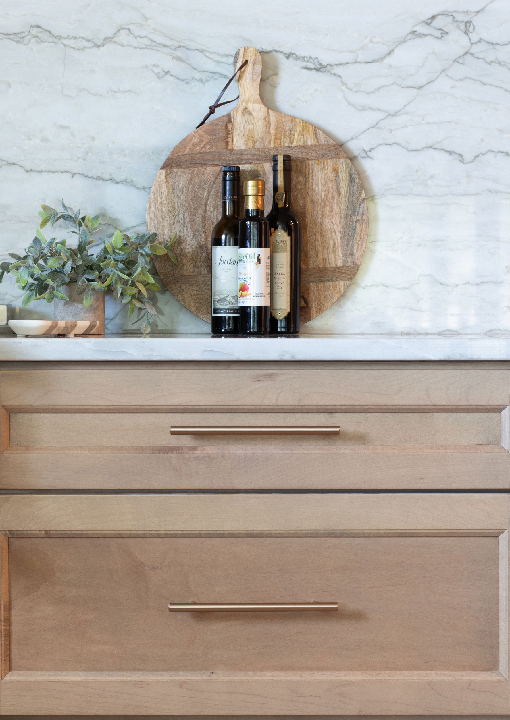 Wood cutting board and olive oil bottles styled on a marble-look kitchen counter with backsplash.