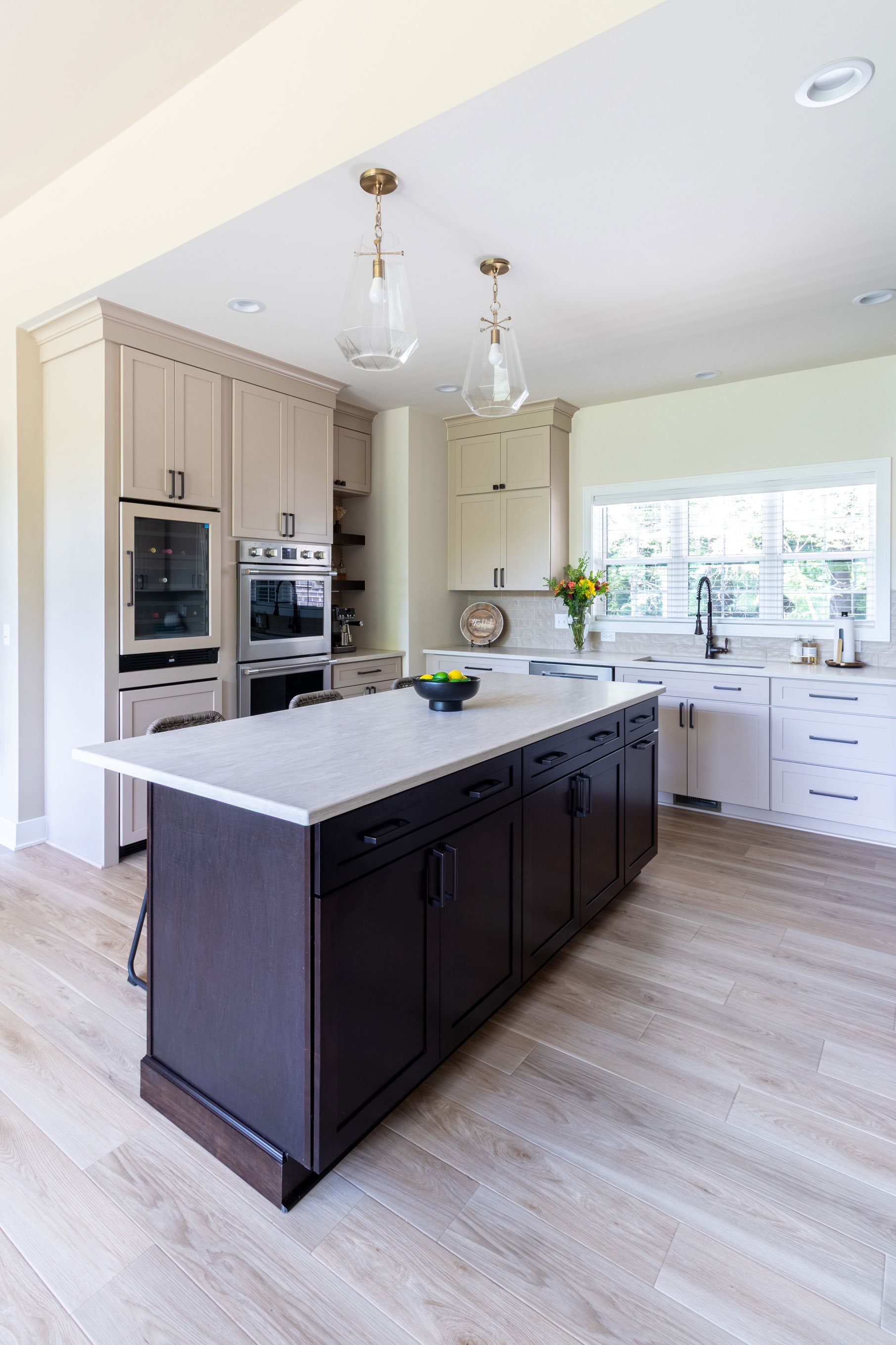 View of the kitchen island in Maple Peppercorn cabinetry alongside the built-in wine refrigerator and double wall ovens.