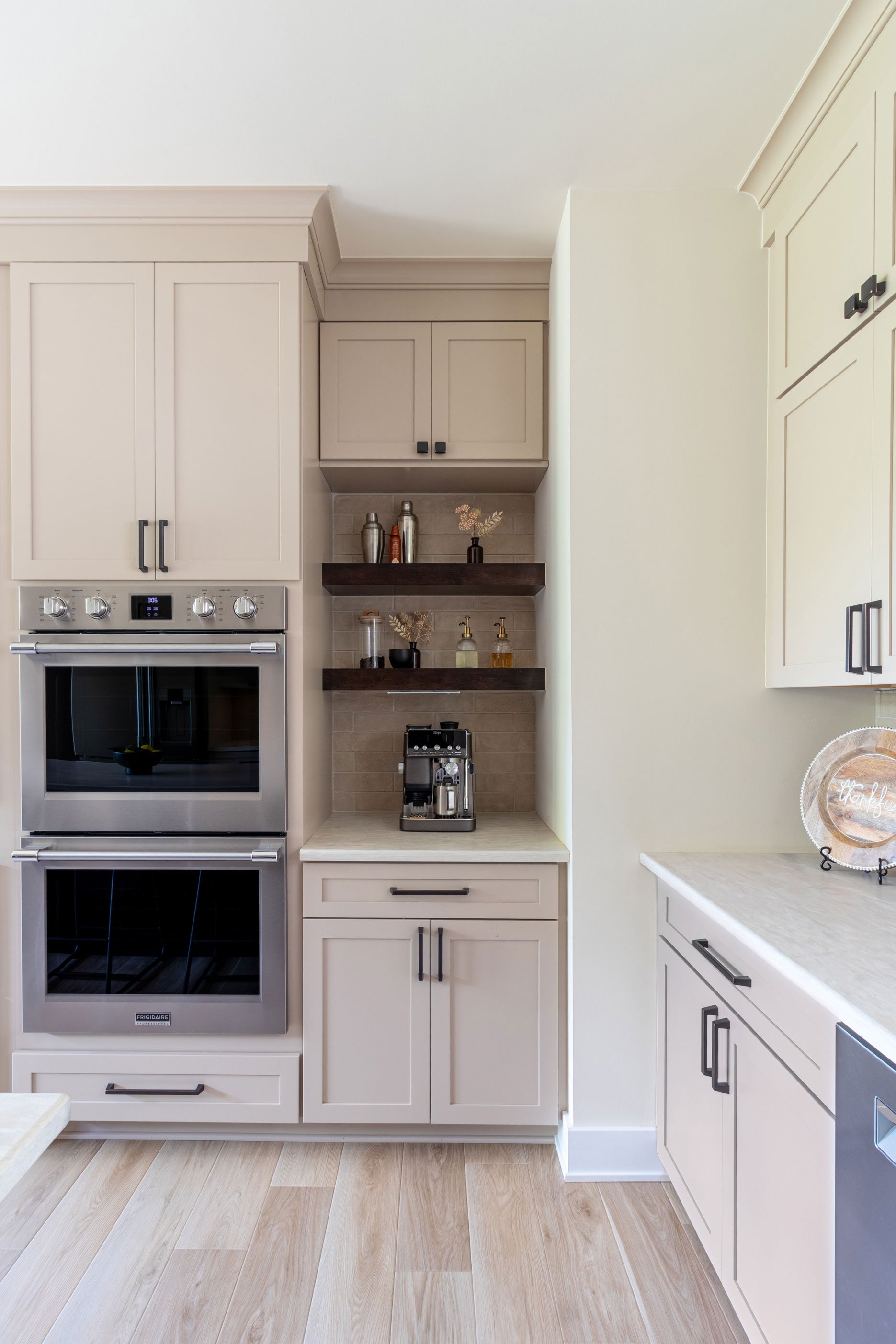 The wall oven and coffee nook area, with Studio Beige cabinetry, floating Maple Peppercorn shelves, and a tucked-in espresso machine.