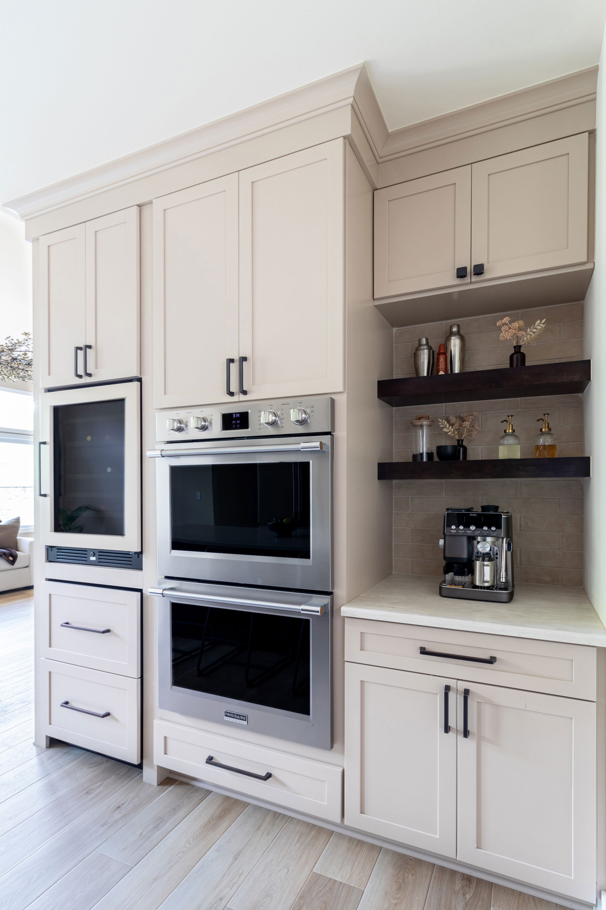 Angled view of the wall oven tower and coffee bar niche with custom panel-front wine refrigerator and floating shelves.