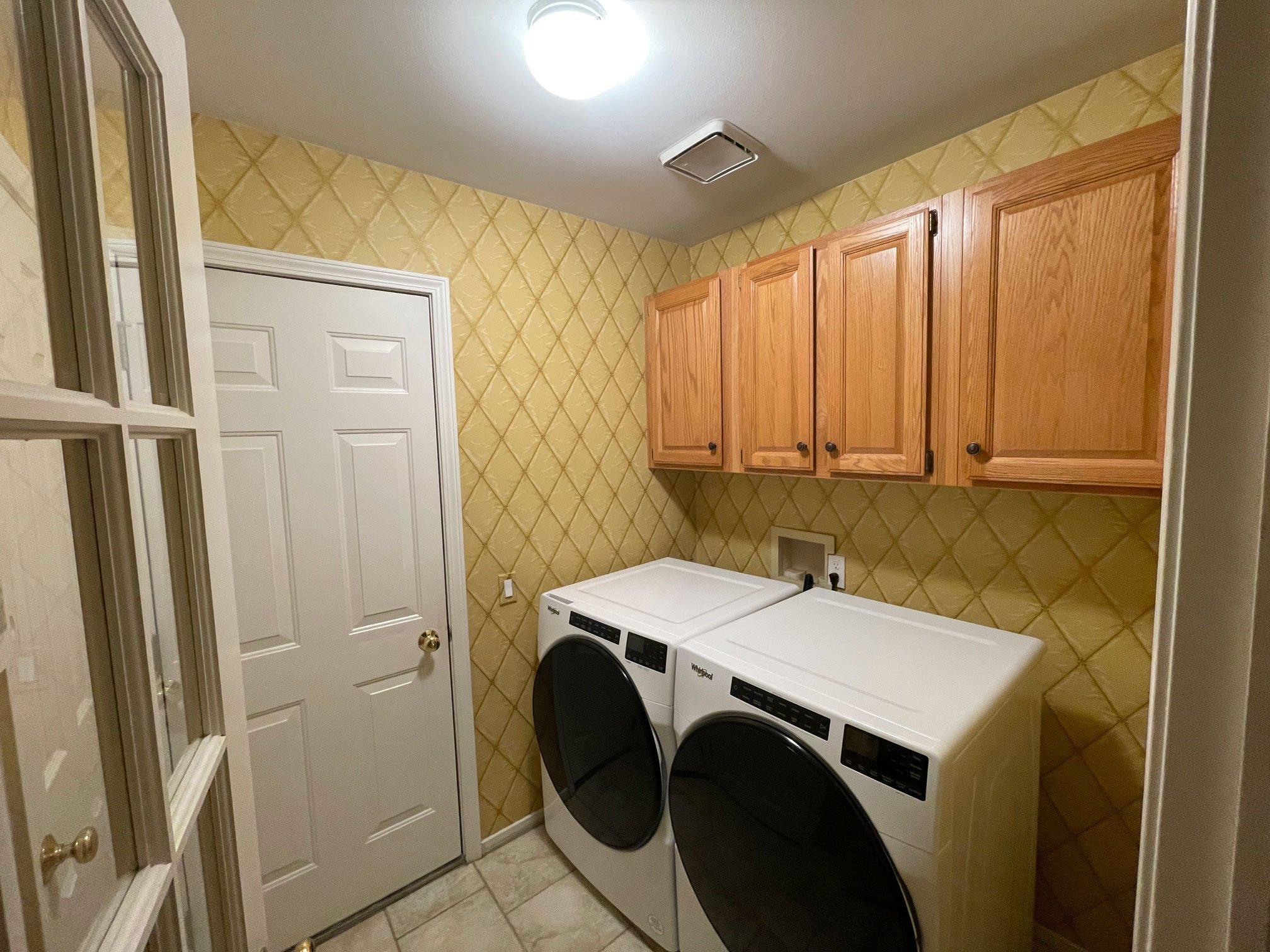 Traditional laundry room with oak wall cabinets, patterned wallpaper, and front-load washer and dryer.
